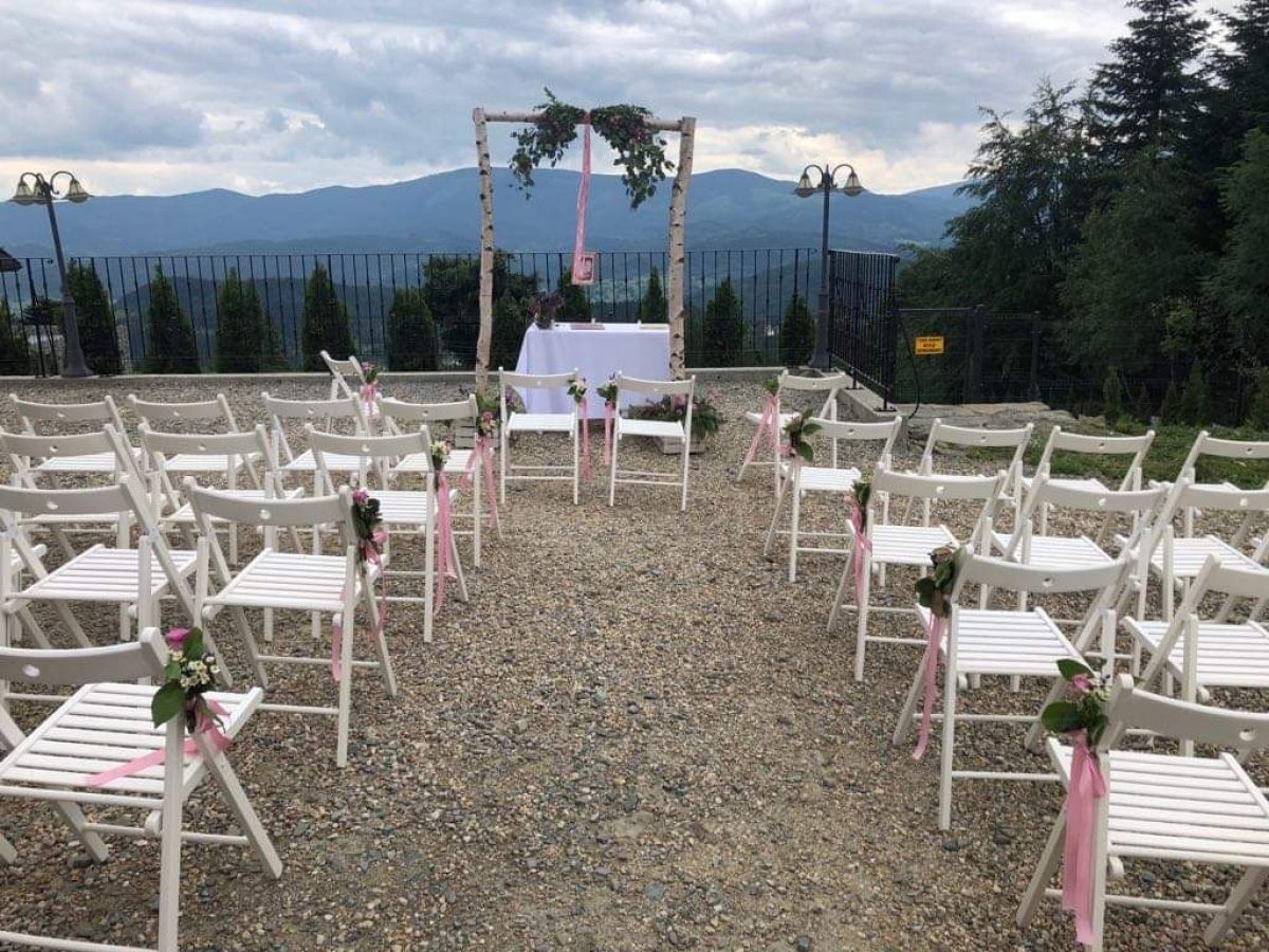 Plenerowa aranżacja ślubna: białe krzesła z różowymi wstążkami i kwiatami, drewniany łuk z dekoracjami z brzozy i kwiatów, stół z białym obrusem, w tle widok na góry i pochmurne niebo.