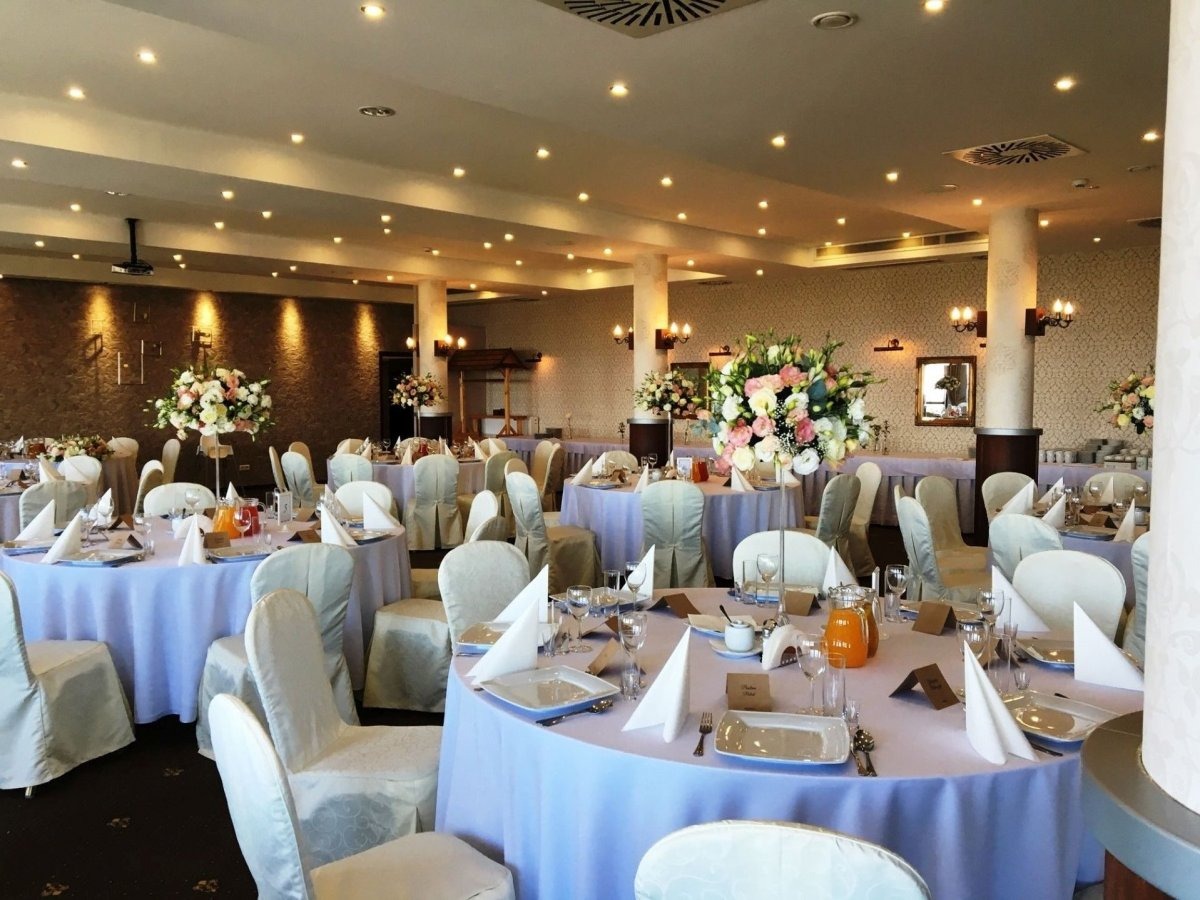 Uroczysta sala balowa z okrągłymi stołami nakrytymi jasnofioletowymi obrusami, białymi krzesłami i kompozycjami kwiatowymi na stołach, przygotowana na przyjęcie.