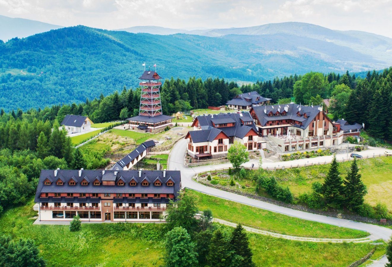 Widok z lotu ptaka na kompleks budynków z wieżą widokową w tle, otoczony zielonymi wzgórzami i lasami.