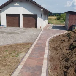 Kaskadowa ścieżka z kostki brukowej w odcieniach czerwieni i beżu prowadząca od garażu do ogrodu, widoczny fragment podjazdu z kruszywa.