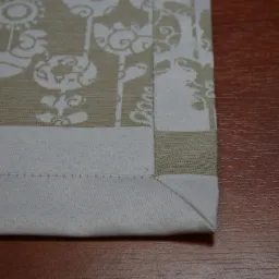 Fragment beżowo-białej serwetki z motywem stylizowanych kogutów i kwiatów, obszytej białą lamówką, leżącej na drewnianym blacie.