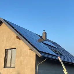 Niedokończony dom z szarą elewacją i panelami słonecznymi na dachu, drabina oparta o ścianę, błękitne niebo