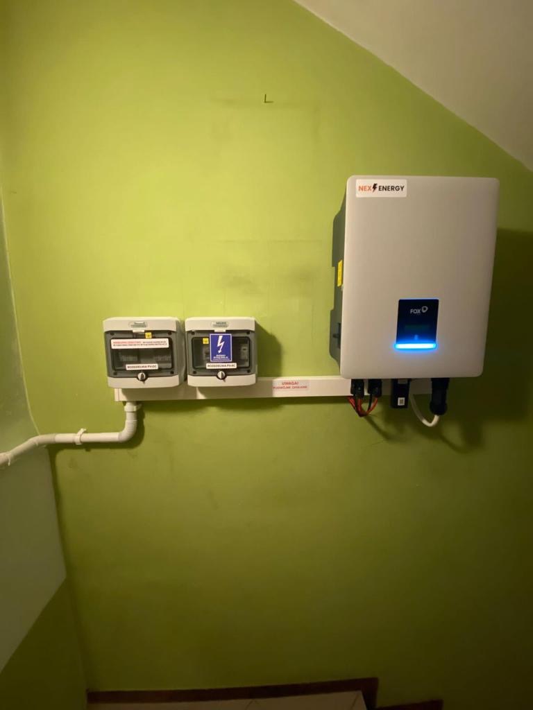 Zamontowany na zielonej ścianie inwerter Nex Energy z podłączonymi kablami oraz dwoma zabezpieczeniami elektrycznymi w białych obudowach.