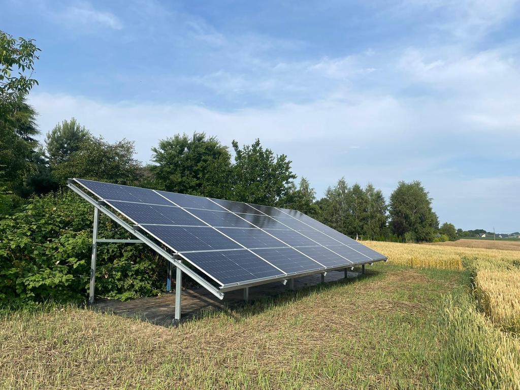 Instalacja fotowoltaiczna na metalowej konstrukcji, umieszczona na skraju pola zboża, z widocznymi drzewami w tle pod błękitnym niebem.