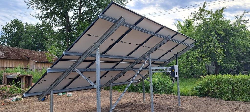 Stelaż pod panele słoneczne zamontowany na gruncie, widok od spodu, z widoczną konstrukcją wsporczą i elementami elektrycznymi, w tle zabudowania gospodarcze i zieleń.