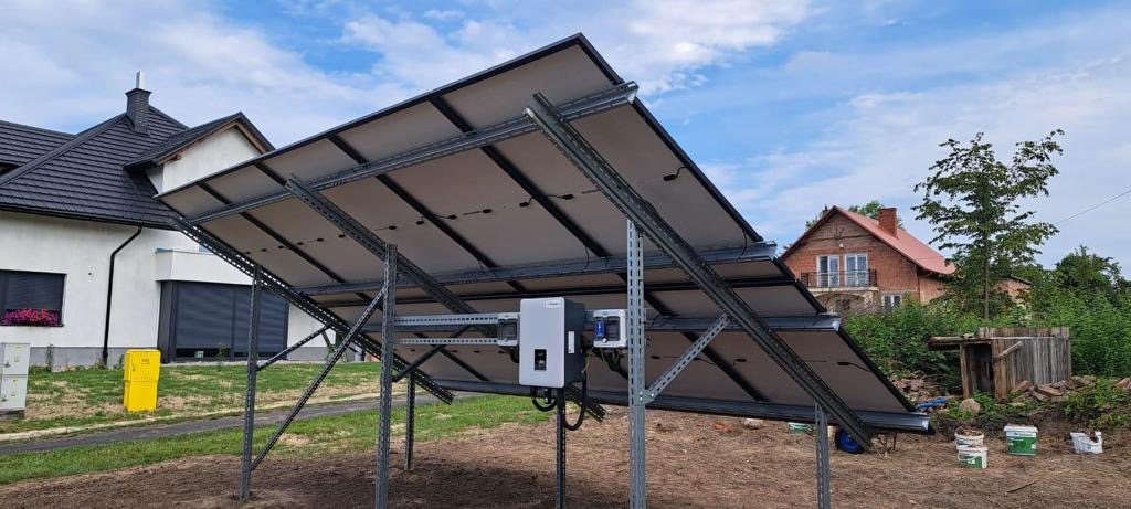Widok od spodu na zamontowane panele słoneczne na konstrukcji wsporczej, z widocznym inwerterem i okablowaniem, na tle dwóch domów jednorodzinnych.
