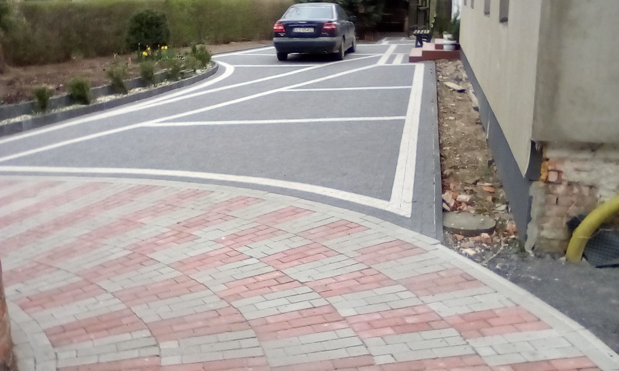 Ułożona nawierzchnia z kostki brukowej w dwóch wzorach: czerwono-szara mozaika i szara z białymi liniami, prowadząca do zaparkowanego samochodu.