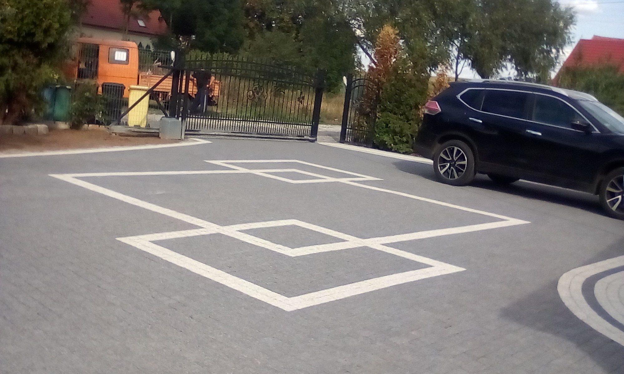 Ułożona kostka brukowa w szarym odcieniu z białym wzorem geometrycznym przed bramą wjazdową i czarnym samochodem osobowym z boku.