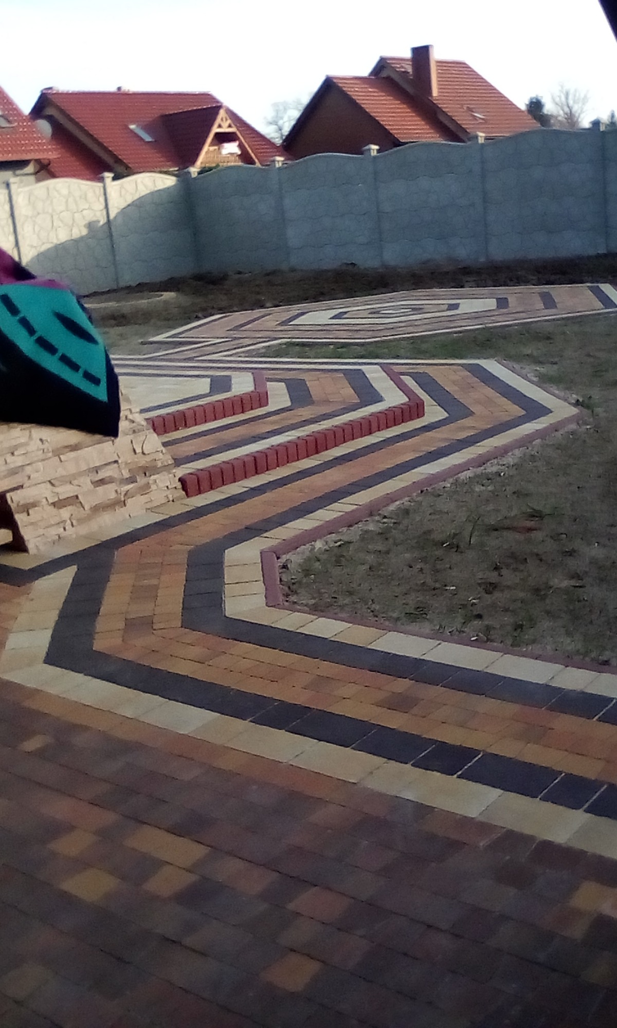 Geometryczny wzór z wielobarwnej kostki brukowej na trawniku przed domem z szarym betonowym ogrodzeniem.