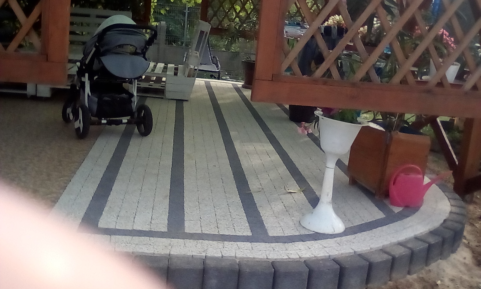 Nowa nawierzchnia z kostki brukowej w odcieniach szarości z ciemnymi pasami, otoczona obrzeżem z ciemnej kostki, na której stoi wózek dziecięcy, biała donica i różowa konewka.