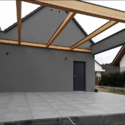 Nowoczesny taras z szarych płytek ceramicznych i drewnianą pergolą, minimalistyczna elewacja w kolorze grafitowym, widoczne oświetlenie zewnętrzne i czarna stolarka drzwiowa