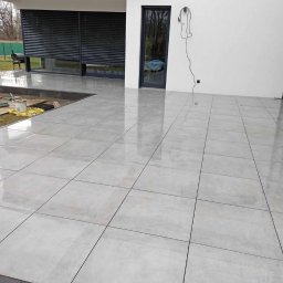 Bonzo - Świeżo ułożone, błyszczące, szare płytki tarasowe na zewnątrz budynku, z widocznymi narzędziami i materiałami budowlanymi w tle, czekające na ostateczne wykończenie.