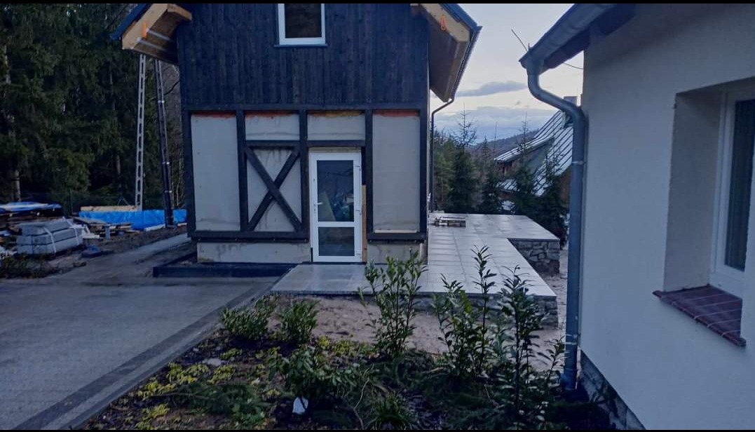 Nowoczesny dom z ciemnego drewna w trakcie wykończenia, widoczny taras z jasnych płyt, kostka brukowa na podjeździe i elementy instalacji odwadniającej.