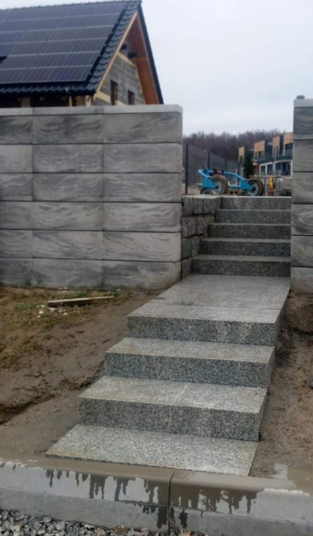 Granitowe schody prowadzące do wejścia, otoczone betonowym murem i widoczną w tle budową domu z panelami słonecznymi.