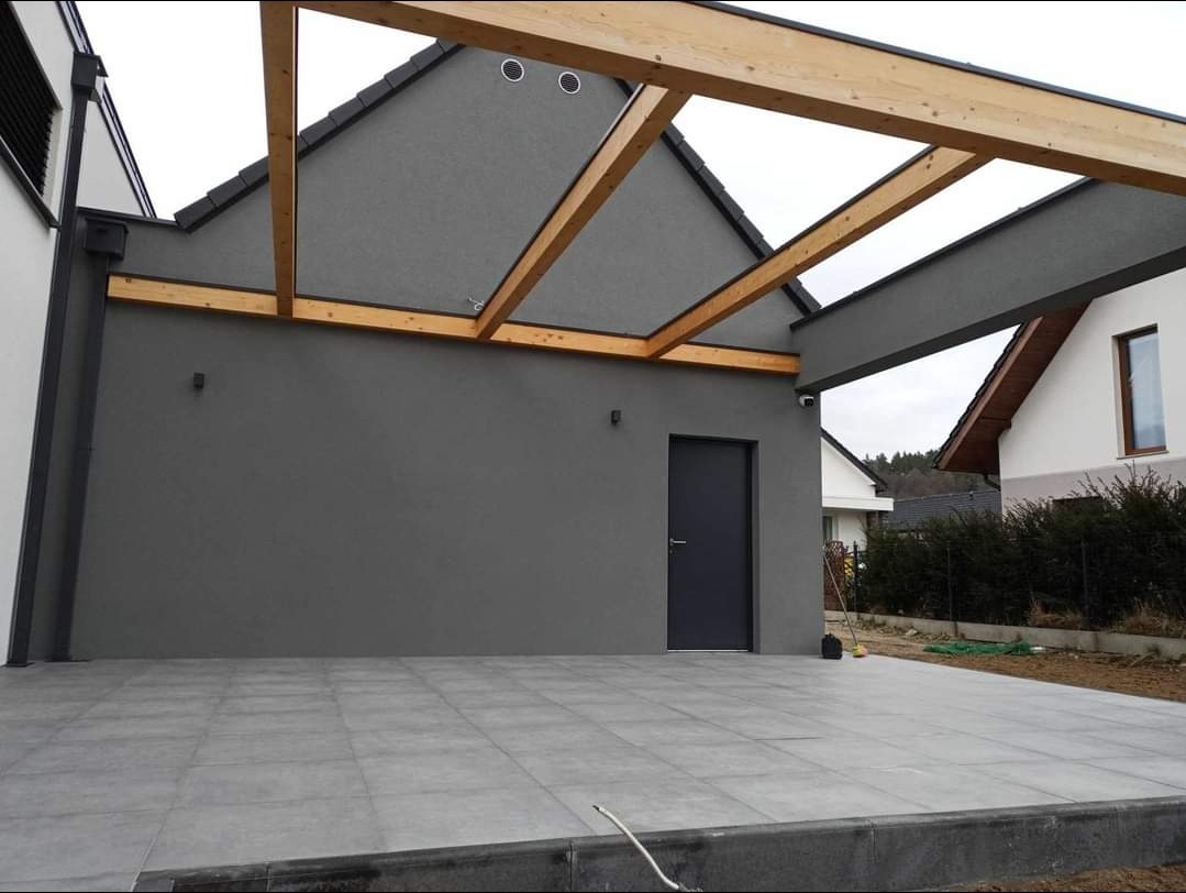 Nowoczesny taras z szarych płytek ceramicznych i drewnianą pergolą, minimalistyczna elewacja w kolorze grafitowym, widoczne oświetlenie zewnętrzne i czarna stolarka drzwiowa