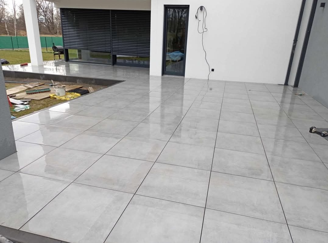 Świeżo ułożone, błyszczące, szare płytki tarasowe na zewnątrz budynku, z widocznymi narzędziami i materiałami budowlanymi w tle, czekające na ostateczne wykończenie.