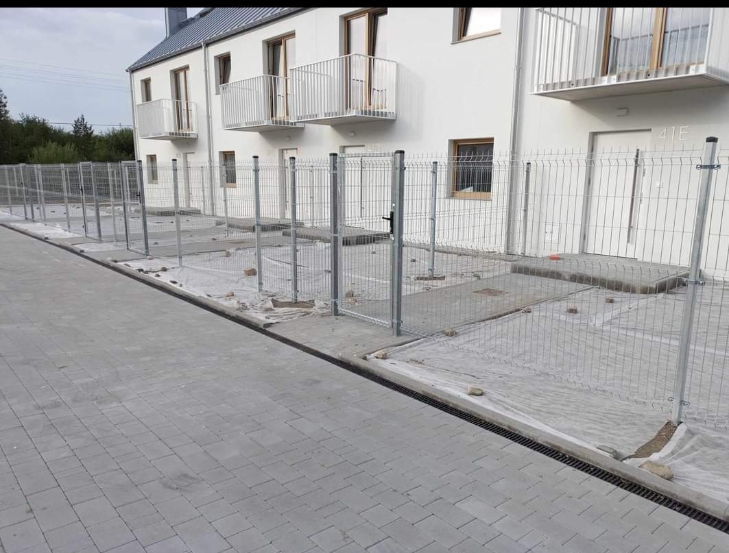 Montaż ogrodzenia panelowego z metalowej siatki przy nowym budynku mieszkalnym z balkonami, widoczne przygotowane podłoże pod trawnik i ułożona kostka brukowa.