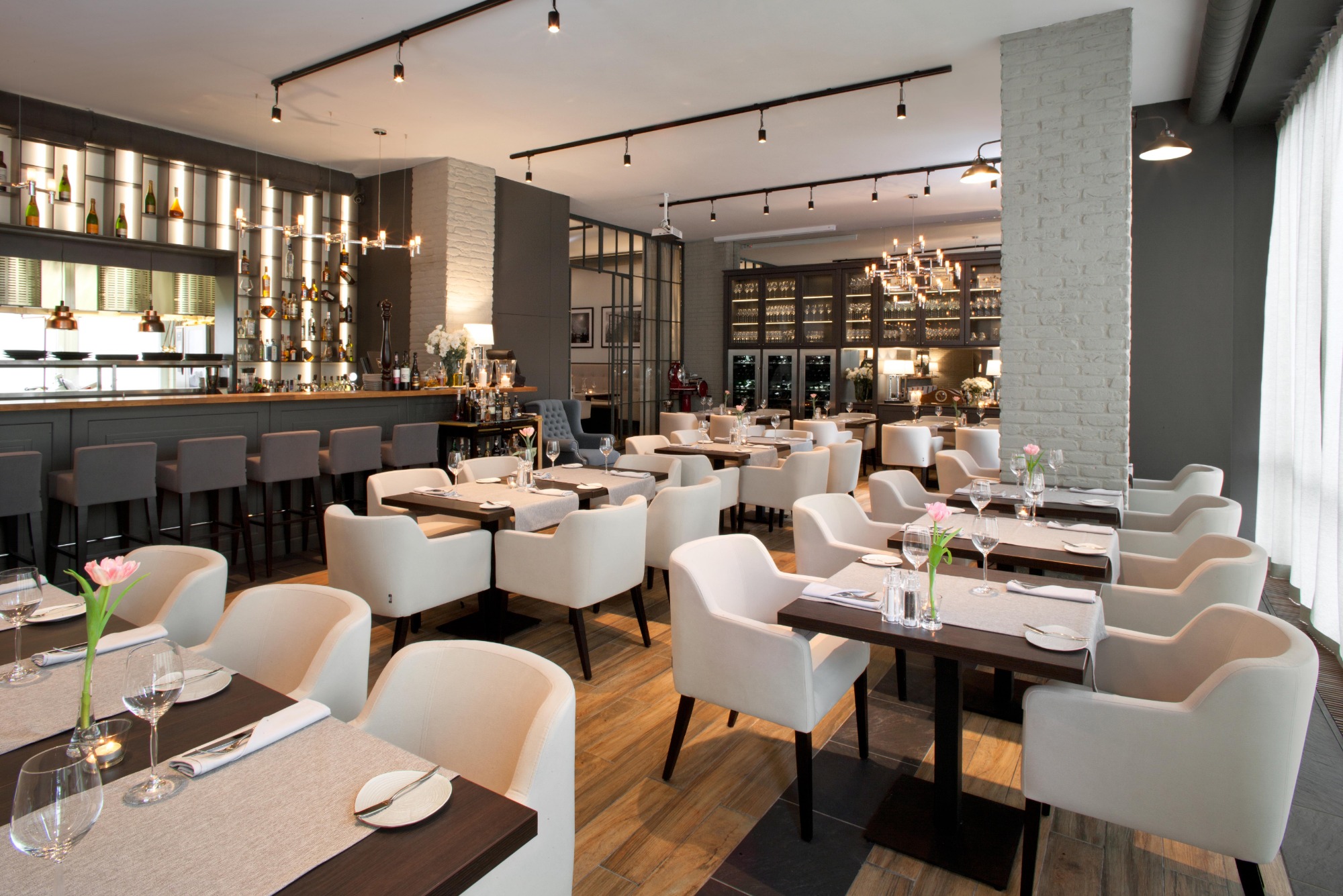 Eleganckie wnętrze restauracji z krzesłami i stołami na wymiar, widoczny bar z podświetlanymi półkami i ekspozycją alkoholi, minimalistyczne dekoracje kwiatowe na stołach.