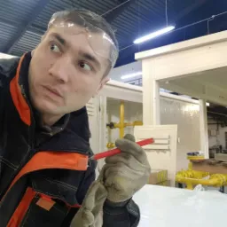 Mężczyzna w okularach ochronnych i roboczych rękawicach, w trakcie pomiaru z użyciem ołówka, na tle instalacji przemysłowej z żółtymi rurami.