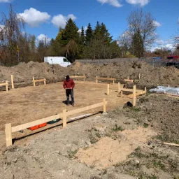 Przygotowanie terenu pod budowę domu jednorodzinnego: wytyczone fundamenty, drewniane szalunki, pryzmy ziemi, pracownik z tabletem na działce.