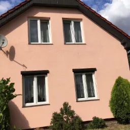 Różowa elewacja domu dwupiętrowego z białymi oknami, brązowymi roletami na parterze i anteną satelitarną po lewej stronie, otoczona zielonymi krzewami.