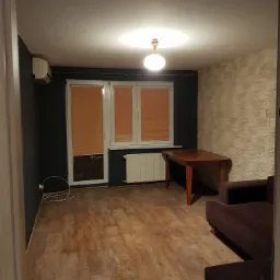 Wnętrze pokoju po remoncie: ciemna ściana z klimatyzatorem, okno z pomarańczowymi roletami, beżowa ściana z imitacją kamienia, drewniany stół i fragment brązowej sofy.