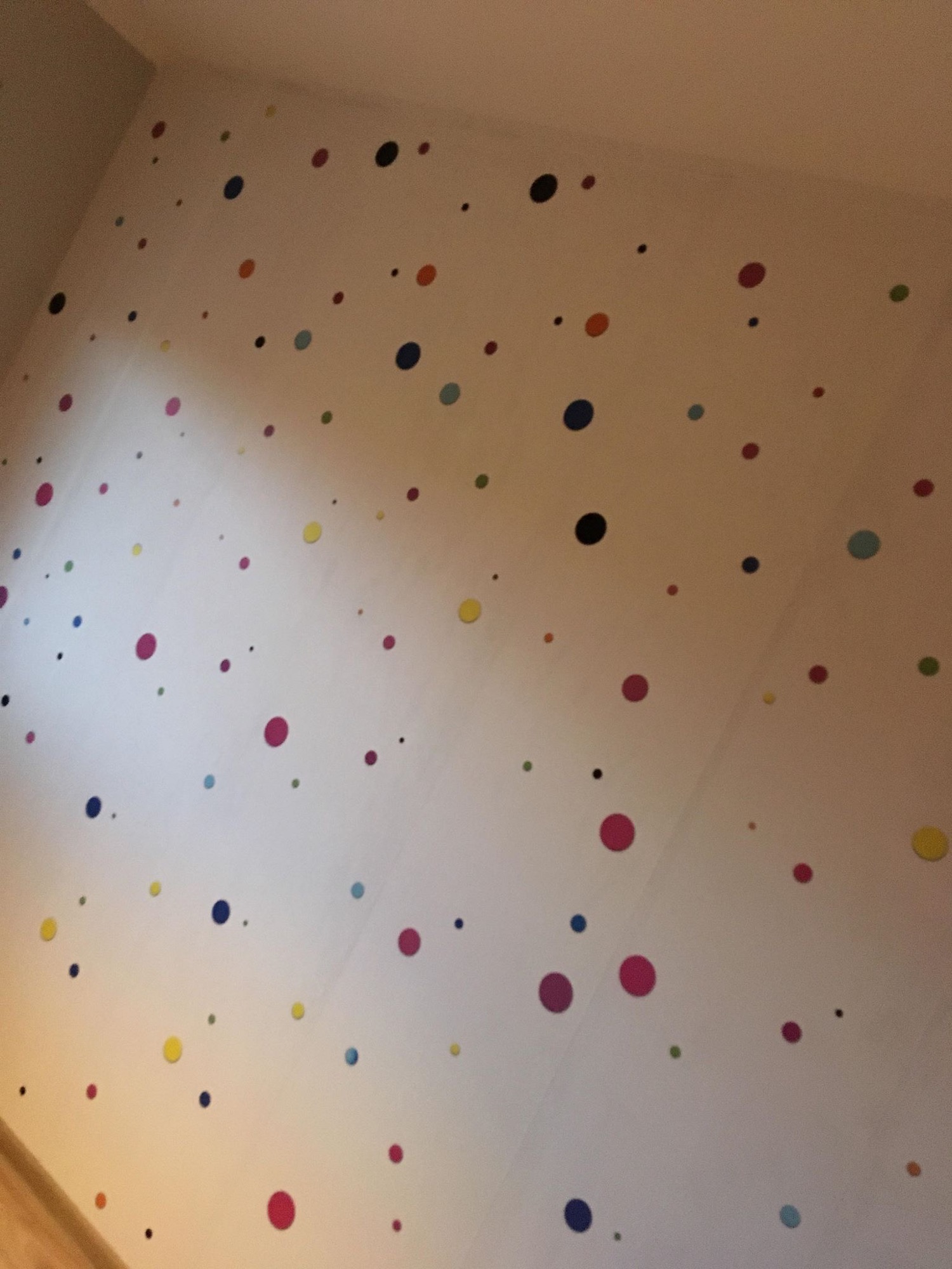 Biała ściana w pokoju pokryta kolorowymi naklejkami w kształcie kropek o różnych rozmiarach i odcieniach, oświetlona z góry.