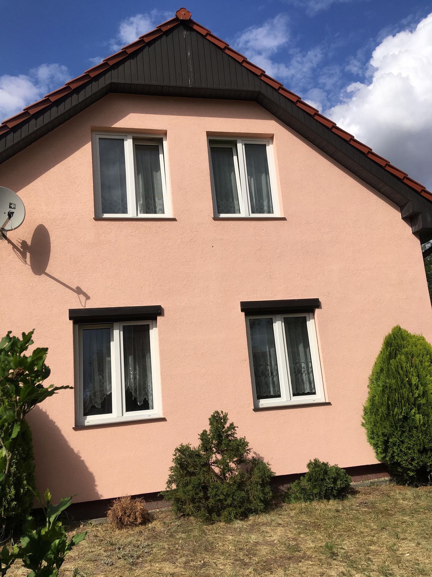 Różowa elewacja domu dwupiętrowego z białymi oknami, brązowymi roletami na parterze i anteną satelitarną po lewej stronie, otoczona zielonymi krzewami.