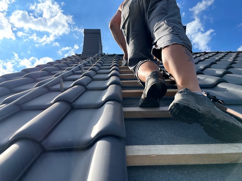 Pracownik w szarych szortach i roboczych butach idzie po drewnianej drabince na dachu z ciemnoszarej dachówki ceramicznej, w tle błękitne niebo z chmurami i komin.