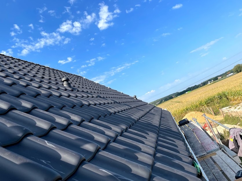 Nowy, czarny dach z ceramicznej dachówki karpiówki, widok z rusztowania na tle błękitnego nieba i pola kukurydzy.