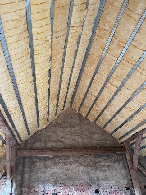 Poddasze w trakcie ocieplania, widoczne metalowe profile i wełna mineralna, surowa ściana z cegieł i drewniane belki konstrukcyjne.