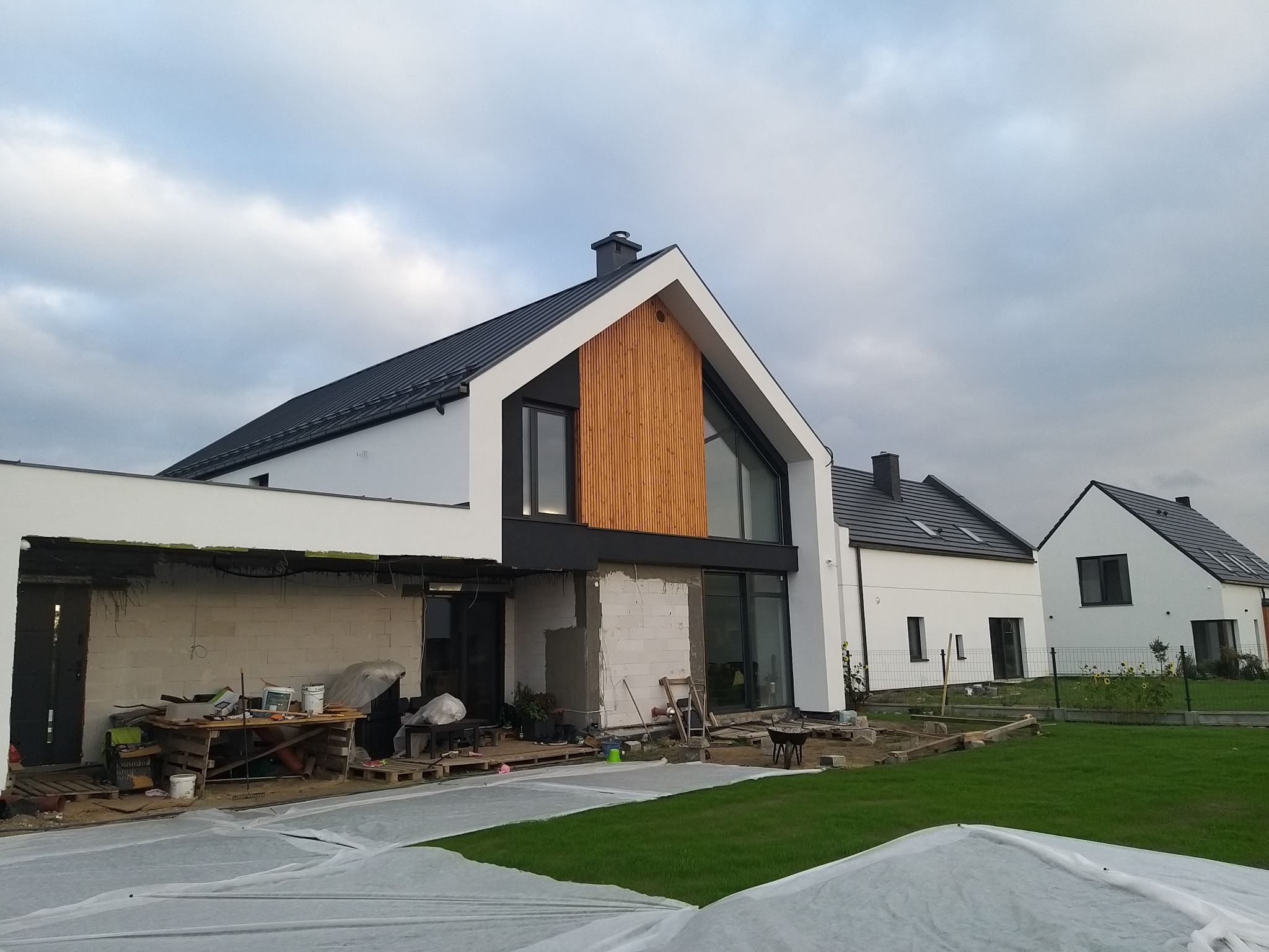 Nowoczesny dom jednorodzinny w trakcie budowy, widoczna surowa elewacja, przygotowane podłoże pod taras, zabezpieczona folią trawa, w tle dwa ukończone domy z czarną dachówką.