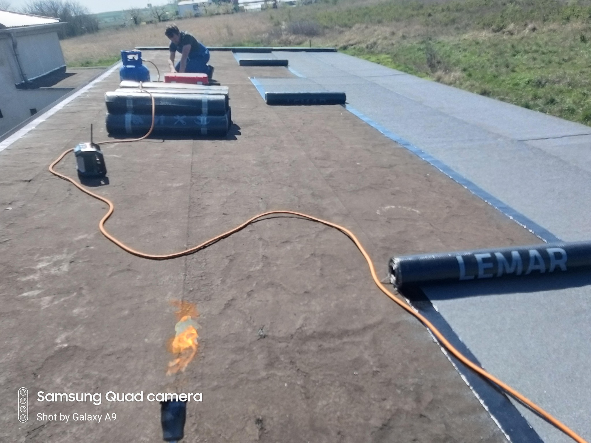 Prace dekarskie na płaskim dachu: dekarz w niebieskim kombinezonie, butla gazowa, palnik i rolka papy z napisem LEMAR. Na dachu widoczne świeżo położone pasy papy. W tle łąka i zabudowania.