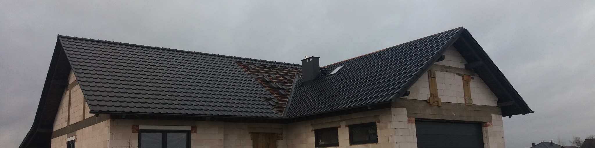 Dom w trakcie krycia dachu, widoczna część z nową, czarną dachówką i fragment z odkrytą więźbą dachową, komin i okno dachowe, ściany z białego pustaka.