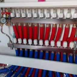 Rozdzielacz ogrzewania podłogowego z czerwonymi i niebieskimi rurami, widoczny manometr i odpowietrzniki, na podłodze folia aluminiowa z siatką.