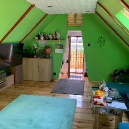 Jasnozielone poddasze w Popowie z drewnianą podłogą i meblami. Sufit wykończony białą, trójwymiarową strukturą. Widoczne elementy dekoracyjne i oświetlenie.