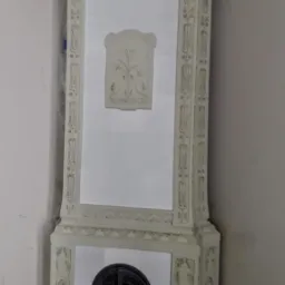 Biały, narożny piec kaflowy z ornamentami roślinnymi i metalowymi drzwiczkami paleniska, umieszczony w rogu pomieszczenia.