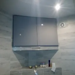 Szare, gładkie szafki wiszące nad blatem z drobnymi przedmiotami, zamontowane na ścianie wyłożonej jasnymi płytkami w łazience lub kuchni.