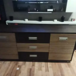 Komoda z frontami w kolorze drewna i czerni, z trzema szufladami i dwojgiem drzwi, na tle podłogi z paneli. Nad komodą półka z dekoracjami.