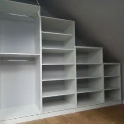 Biała zabudowa w skosie poddasza z drążkami na ubrania i półkami, widok z dołu.