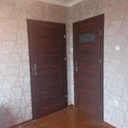 Dwa brązowe, drewniane drzwi wewnętrzne na tle ściany z geometryczną tapetą w odcieniach szarości i różu, widoczne gniazdko elektryczne i włącznik światła.