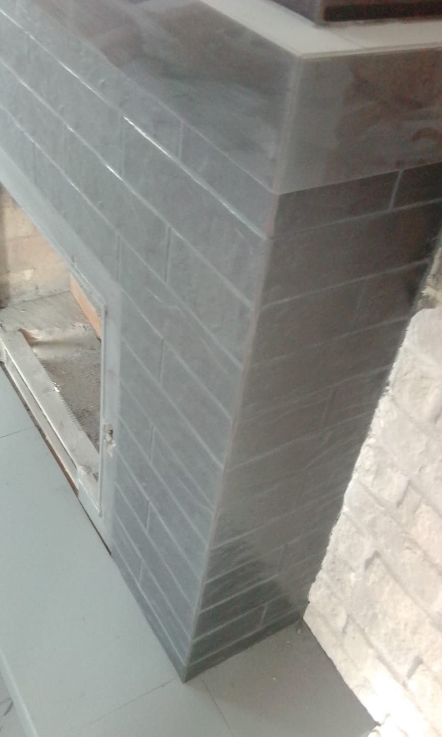 Fragment obudowy kominka wyłożonej szarymi płytkami imitującymi cegłę, widoczne niedociągnięcia w fugowaniu.