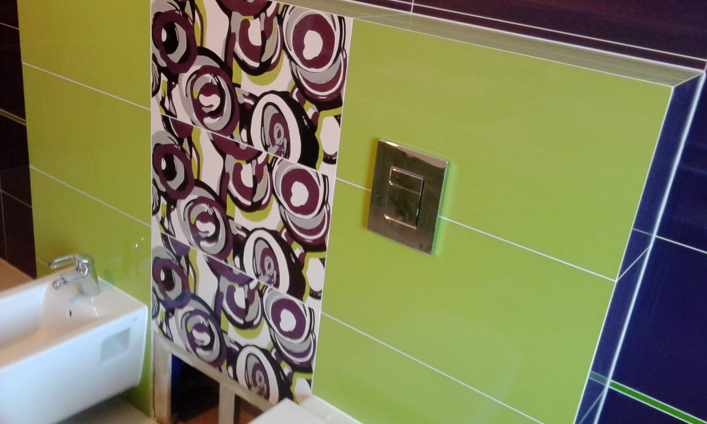 Ściana łazienki wyłożona zielonymi płytkami z poziomymi fugami, obok panel z płytkami z wzorem kół, chromowany przycisk spłuczki i fragment umywalki.