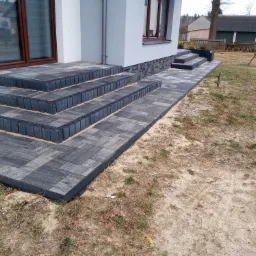 Nowoczesne schody wejściowe do domu, wyłożone szarą kostką brukową i obrzeżami, z widocznym fragmentem elewacji budynku w stonowanej kolorystyce.