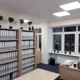 Wnętrze biura z regałami wypełnionymi szarymi segregatorami, małymi drzewkami w doniczkach na górze regałów i na parapecie, biurkiem z komputerem i drukarką przy oknie.