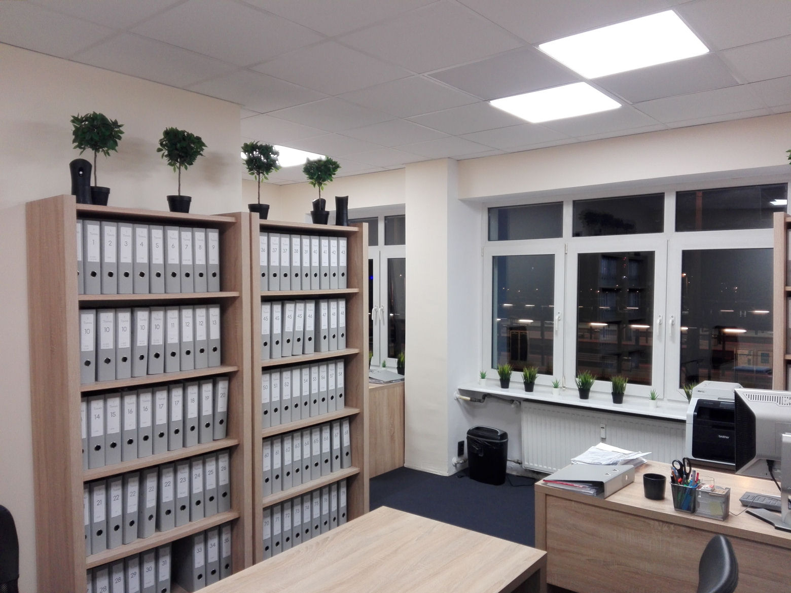 Wnętrze biura z regałami wypełnionymi szarymi segregatorami, małymi drzewkami w doniczkach na górze regałów i na parapecie, biurkiem z komputerem i drukarką przy oknie.
