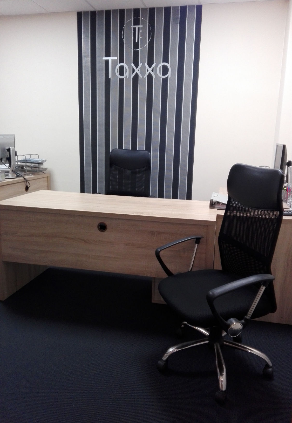 Nowoczesne biuro: jasne biurko z drewnianym wzorem, czarne krzesło biurowe z siatkowym oparciem na ciemnej wykładzinie, w tle logo 'Taxxo' na ścianie z szarymi pasami.