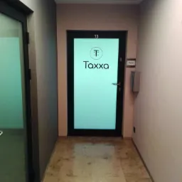 Korytarz biurowy z drzwiami do biura rachunkowego Taxxa, numer 13 nad drzwiami, tabliczka z nazwą firmy na ścianie, marmurowa podłoga.