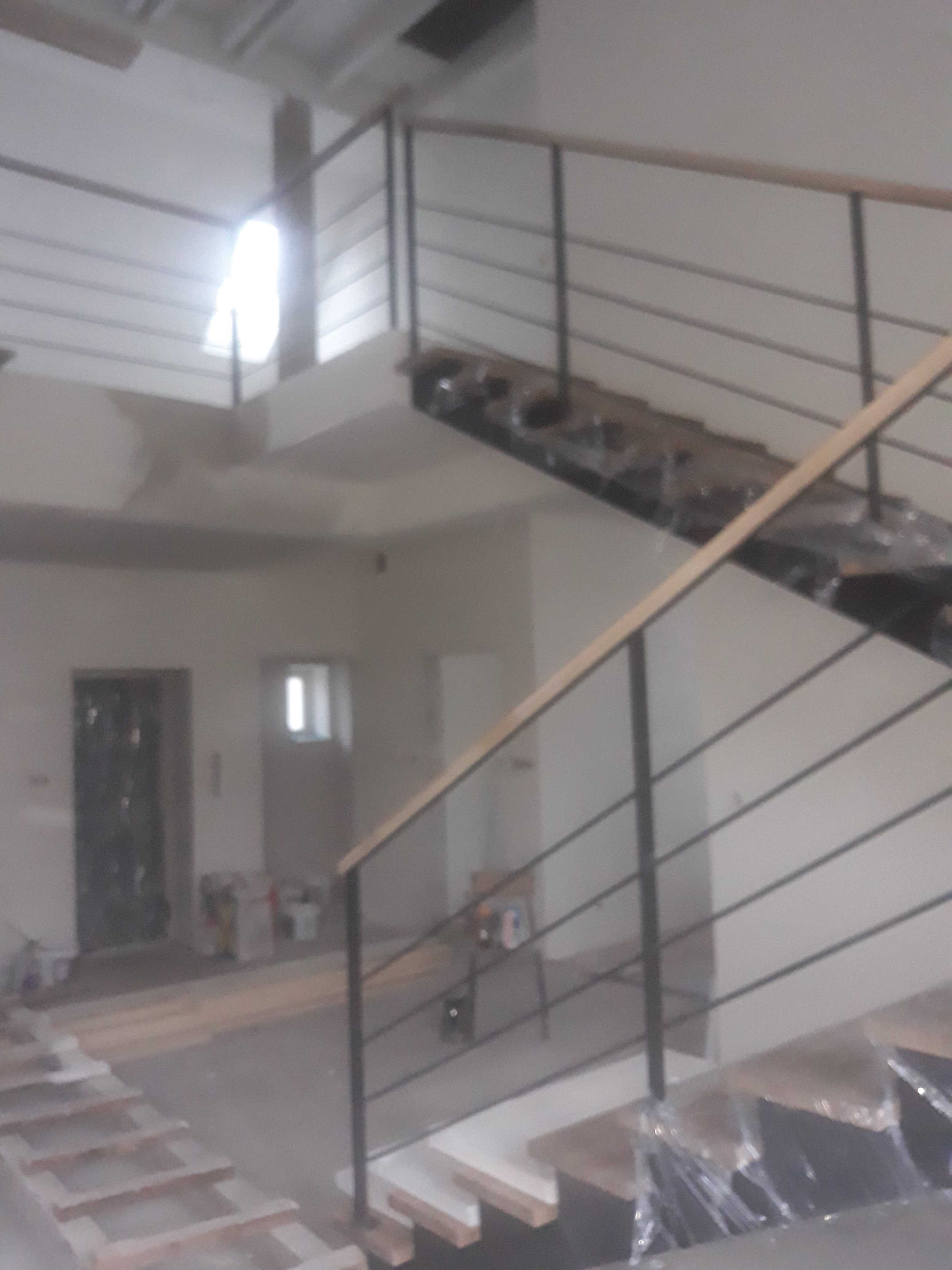 Wnętrze w trakcie remontu: widoczne schody z metalową balustradą i drewnianymi poręczami, częściowo zabezpieczone folią. W tle surowe ściany, okno i drzwi, a także materiały budowlane.