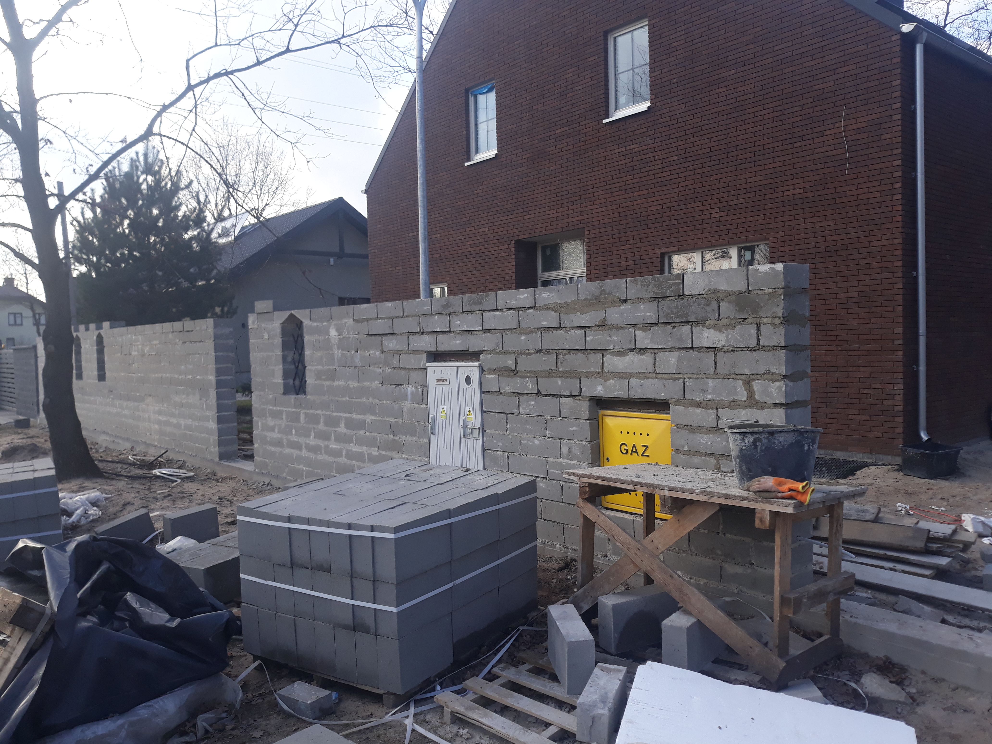 Wznoszenie muru z szarego bloczka betonowego na posesji, widoczna skrzynka gazowa w ścianie, paleta z bloczkami, wiadro i robocze rękawice na drewnianym podeście.